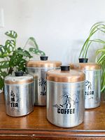 Four Hawthorn aluminum canisters labeled Flour, Sugar, Coffee, and Tea with copper-colored lids and black knob handles on a wooden surface with plants in the background. Shows wear including fading and dents.