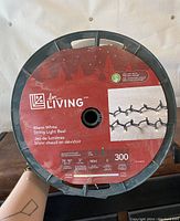 Photo of one reel with label showing brand 'For Living', product details including warm white string light, 75' 10" length, 300 mini LED bulbs, 3 inch spacing, cool proof, and outdoor rated.