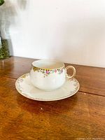White tea cup and saucer set displayed on wooden surface, showing gold rim and floral design on cup and saucer.