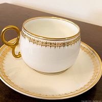 Single teacup and saucer showing gold rim, handle, and detailed gold decorative pattern on ivory bone china.