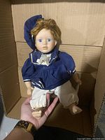 Front view of porcelain boy doll wearing navy sailor jacket and white pants inside a cardboard box, held in hand to show size and features.
