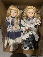 Two porcelain dolls standing side by side in a cardboard box; one wears dark blue dress with white lace, the other in a blue-checkered outfit with a red rose and lace collar, both with blonde hair and pale complexions.