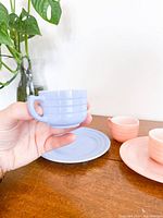 Blue cup held in left hand with pink cup and saucer, pink creamer, and pink saucer next to it on wood surface with vase plant in background.