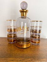 Full shot of whisky decanter with stopper alongside four matching glasses showing gold rim and purple hue.