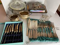 Full lot overview showing boxed antler handled steak knives, partial cutlery set, and silver plated serving dishes on table.