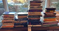 Photo showing multiple stacks of various hard and soft cover books arranged on a couch by a window.