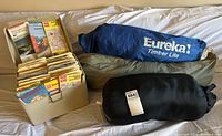 Blue Eureka Timber Lite tent bag, green tent bag, black sleeping bag sack and box of maps on white surface.