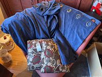 Three blue corduroy curtain panels with large silver-tone grommets draped over a chair, alongside two multicolored abstract pattern throw pillows.