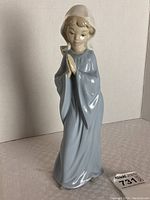 Full view of the Nao porcelain figurine of the child in a blue robe standing on a white surface against a plain background.