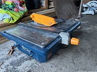 Side view of Ryobi wet tile table top saw showing blue metal body, silver cutting surface with measurement guides, and orange blade guard.