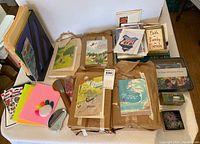 Top view of a large collection of greeting cards, four brown kraft paper-wrapped bundles of cards, colorful sheets of paper, markers, and office supplies arranged on a floor with a white wall backdrop.