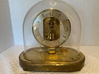 Front view photo of vintage anniversary clock housed in clear glass dome on brass-tone oval base, showing clock face with gold-tone numerals and pendulum.
