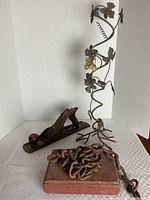Wide side view showing wooden hand planer, coiled heavy chain on metal base, and tall decorative wrought iron rack with grapevine design