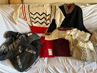 Lot of seven women’s sweaters laid out on white fabric backdrop showing variations in color and style including a prominent white sweater with red and navy zigzag design and solid red waistband, a black V-neck with trim, and additional sweaters in cream, burgundy and grey shades.