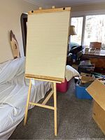 Wooden tripod easel standing upright with a large attached notepad displayed on it. Background shows white covered couch and boxes.