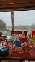Table displaying various coloured glass serving dishes including red, aqua blue, ruby, amber pieces arranged near a window with outdoor view.