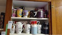 Two shelves of assorted coffee and tea cups and mugs shown inside a white cabinet, various decorative and functional styles.