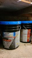 Two buckets side by side under shelf showing labels and lids