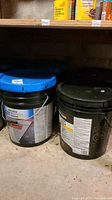 Two 17L buckets of driveway sealer placed on concrete floor; left is Rust-Oleum with blue lid, right is Armor Coat with black lid.