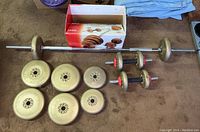 Vinyl dumbbell plates, two dumbbell bars with red collars, one long barbell bar, and original box on carpeted floor.
