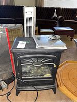 Electric stove with black metal body, glass front panel showing faux logs, power cord visible, with a tower fan and thermo clock in background.