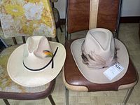 Two hats displayed on chairs: a beige pure wool feathered urban hat and a light beige woven hat with a black band and colorful feather.