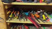Wooden shelf with multiple sets of pliers of various types and sizes with different colored handles including yellow, red, orange, blue and black lined up.