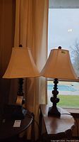 Photo showing both matching table lamps placed side by side in front of a window with beige square shades and dark turned wood bases.