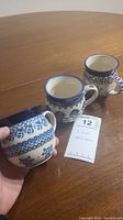 Three ceramic cups placed on a wooden table showing blue floral patterns and a tag.