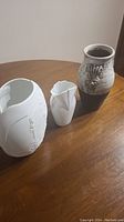 Photo of three vases on a wooden table including a large white ceramic vase, a smaller white geometric design vase, and a stoneware vase with glaze.