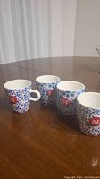 Four ceramic coffee cups with floral design and red DE badge logo on wooden table