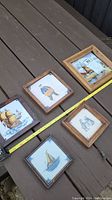 Five Dutch antique ceramic tiles in wooden frames arranged on wood surface, showing various traditional Dutch scenes including ships, people, and a windmill.