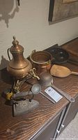 Photo showing assortment of antique items including brass kettle, cast iron flat iron, wooden scoop, cast metal pot, and black cast iron pan in background
