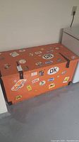 Front and side view of the large orange toy box with multiple stickers and black metal hinges.