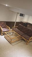 Overall view of three-piece rattan wicker furniture set including sofa, armchair and glass-top coffee table in basement setting.