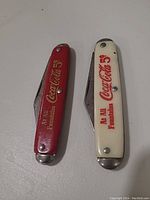 Two Coca-Cola 5¢ branded pocket knives, red and white handle, with 'At All Fountains' text.