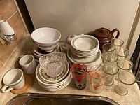 Photo showing stacked dishes including white bowls and plates along with a brown teapot, various clear glasses, and vintage printed glasses