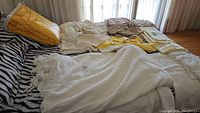 Wide view of assorted handmade crocheted blankets and towels laid out on a bed, including a large white fringed blanket and others in yellow, brown, and beige