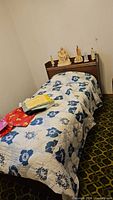 Full view of Captain's bed with headboard against the wall, covered with a white quilt with blue floral print. Various small figurines are placed on the headboard shelf.