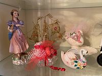 Collection of assorted ceramic figurines: standing woman figurine, sailing ship centerpiece, swans with ribbons and netting, displayed on glass shelf.