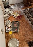 Wide shot showing assortment of ceramic plates, bowls, mugs, glass and plastic trays on floor along with various glassware stacked near corner