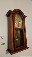 Full view of the wooden wall clock hung on wall showing the entire clock case and pendulum inside glass panel.