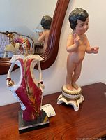 Photo of two figurines including a brightly colored Porcelanas female figure and a religious child figure standing on a table in front of a mirror.