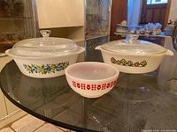 Set of three vintage glass bake dishes on glass table: Fire King with blue flowers and green leaves, Glasbake with orange floral design, and smaller white dish with red geometric pattern.