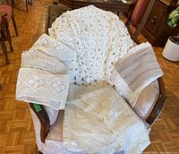 Wide view of all crocheted textiles displayed on a pink accent chair, showing folded pieces and a large table cloth draped over the back.