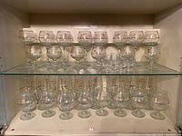 Wide shot of all 33 glassware items displayed on three shelves including 5 styles of stemmed glasses and 4 shot glasses, all with gold Olympic logos and detailing.