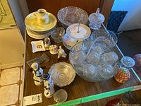 Overview of lot items including glass punch bowl with cups, tiered plate, ceramic plates, figurines, and some glass dishes on wooden table.