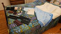 Wide shot of all items on a bed covered with a colorful patterned bedspread including multiple purses, wallets, linens, and pajamas package.