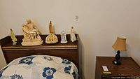 Religious ceramic figures including Madonna, Mary, Jesus, and others displayed on the bed headboard with wood finish. Figures vary in size, some appear beige, yellow, and white.