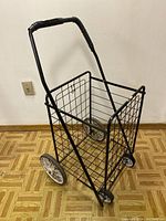 Side view of black metal wire shopping cart with large rear wheels and smaller front wheels, showing the curved handle with plastic grip.